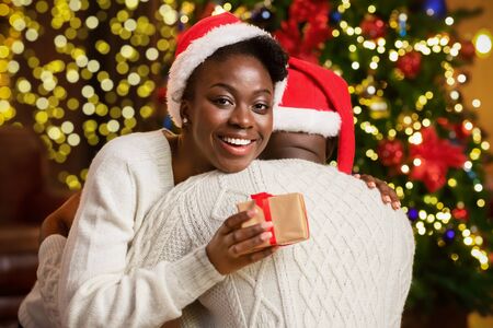 Happy black girl embracing her boyfriend, grateful for lovely christmas present, holding gift box and looking at cameraの写真素材