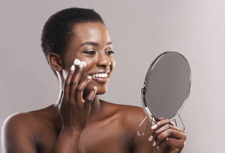 Skin care concept. Beautiful African American Woman Looking in Magnifying Mirror And Applying Face Cream Over Gray Studio Backgroundの写真素材