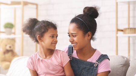 Happy african american sisters smiling to each other, sitting at home, panorama
