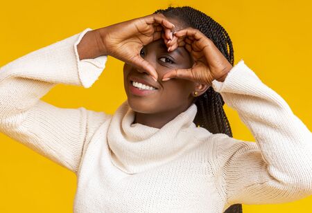 Smiling loving black winter woman shaping hands like heart over her face, yellow background
