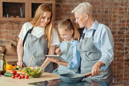 New cooking app. Little girl showing mom and granny new recipes on digital tablet, kitchen interiorの写真素材