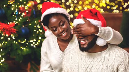 Time for presents. Loving black wife closing husbands eyes with hand, prepared Christmas surprise for him.の写真素材