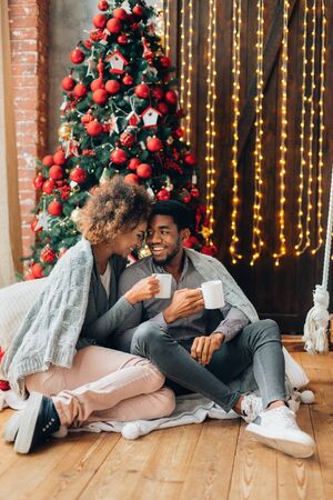 First winter together. Loving african-american couple enjoying Christmas morning coffee, sitting near decorated tree at home, copy spaceの写真素材