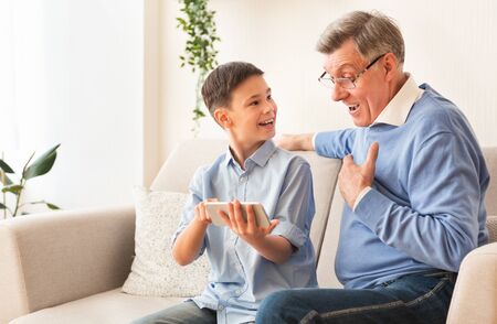 Smiling Boy Showing Grandfather How To Play Mobile Games On Smartphone Sitting On Sofa Indoor. Copy Spaceの写真素材