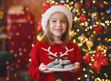 Cheerful little girl in Santa hat holding plate with chocolate cookies Santa Claus