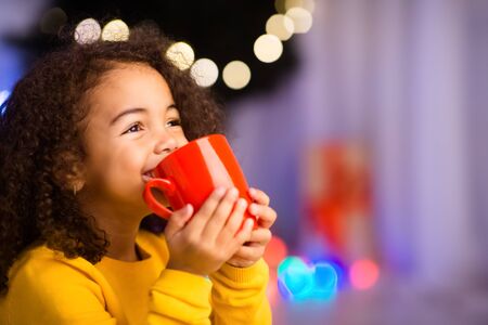 Cute little african girl with cup of hot cocoa sitting near Christmas tree, empty spaceの写真素材