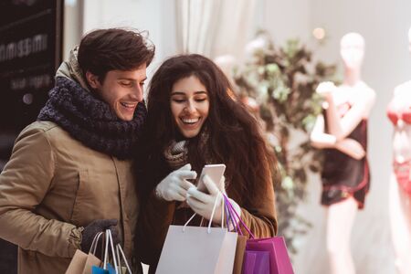 Happy Spouses Shopping And Using Phone Walking In City In Winter Night. Discount Finder, Mobile App Concept.の写真素材