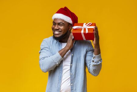 Guess whats inside. Happy black man in Santa hat shaking Christmas present, listening what rattles in gift box, yellow backgroundの写真素材
