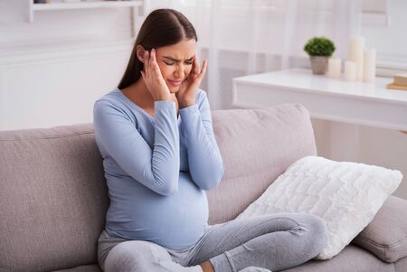Headache During Pregnancy. Expectant Woman Having Severe Migraine Massaging Temples Sitting On Sofa...