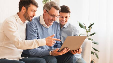 Elderly Man Using Laptop Computer Sitting Between Middle-Aged Son And Grandson On Sofa Indoor....