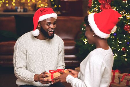 Romantic black couple in Santa hats presenting gifts to each other while celebrating christmas at homeの写真素材