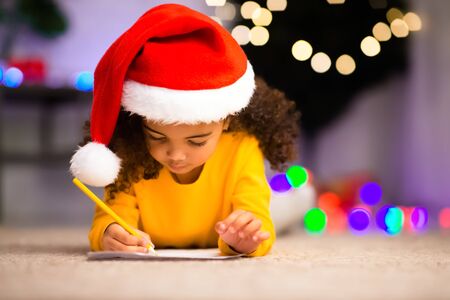 Cute black girl in Santa hat writing presents list for parents on winter holidays, free spaceの写真素材