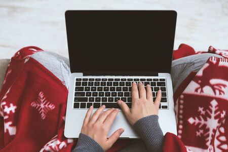 Christmas advertisement. Woman covered by red plaid using laptop with black screen at home, copy spaceの写真素材