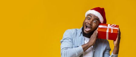 Funny african american guy holding Cristmas gift box and wonder whats inside, yellow background, panoramaの写真素材