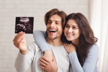 Happy young couple holding and looking at ultrasound scan of their baby, cuddling and bonding at homeの写真素材