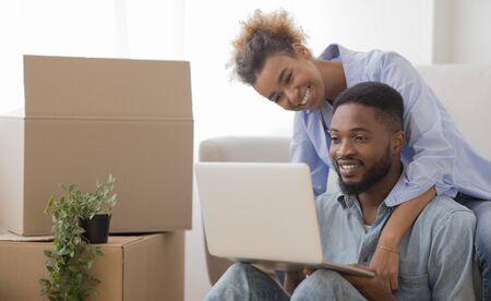 Positive African American Couple Using Laptop Sitting On Couch Among Moving Boxes In New Apartment. Selective Focusの写真素材