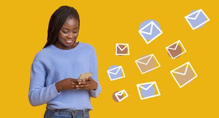 African woman typing text message on smartphone, e-mail inbox envelopes flying from phone, yellow background, panoramaの写真素材