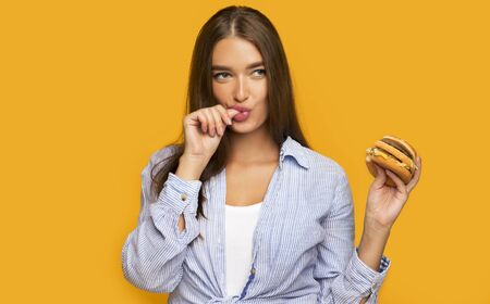 Hungry Girl Holding Burger Licking Fingers Standing Over Yellow Studio Background.の写真素材