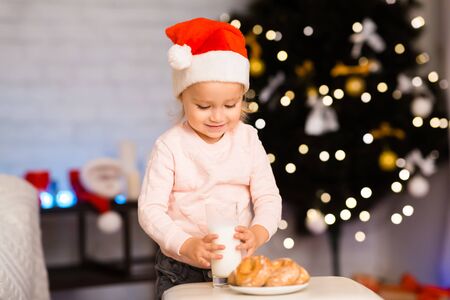 Little baby girl preparing milk and cookies for Santa Claus near Christmas treeの写真素材