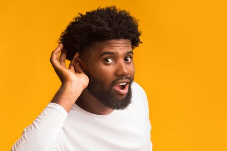 Curious interested african american man holding hand near ear and trying to hear information, orange studio background, free spaceの写真素材