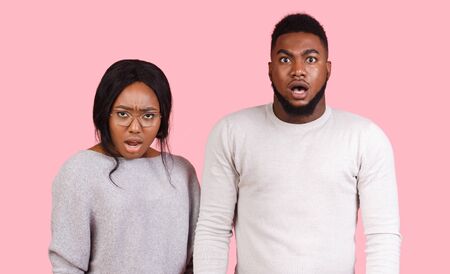 Amazed african man and woman with open mouths looking at camera over pink backgroundの写真素材