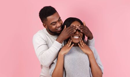 Black happy man making surprise for girlfriend over pink background, covering girl eyes with palms, panorama with copy spaceの写真素材