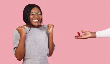 Extremely happy black girl looking at engagement ring in man hand, pink backgroundの写真素材