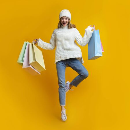 Emotional young blonde woman in woolen hat jumping with shopping bags over yellow background