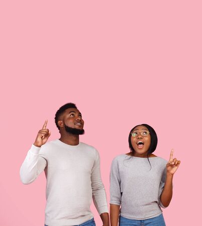 Amazed african american man and woman pointing fingers upwards and looking at free space on pink studio backgroundの写真素材