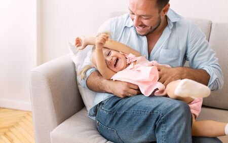 Best Dad In The World. Millennial man tickling his daughter who lying on his lap on the sofa. Free spaceの写真素材