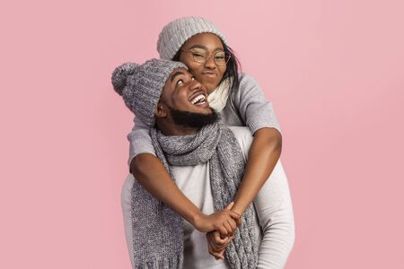 Afro couple in love wearing winter clothes, cuddling over pink background, girl hugging guy from...