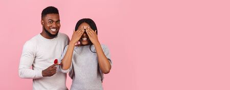 Amazing surprise on St Valentines Day. Romantic black man presenting his girlfriend beautiful ring over pink background, girl covering eyes with palms, panorama with copy spaceの写真素材