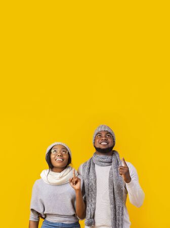 Unbelievable sale. Interested african american winter man and woman pointing fingers upwards and looking at free space on yellow studio backgroundの写真素材