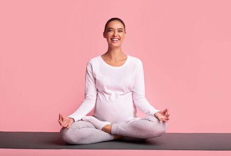 Pregnancy Meditation. Smiling Pregnant Girl Doing Yoga Sitting In Lotus Pose On Gymnastics Mat Over Pink Background. Studio Shotの写真素材