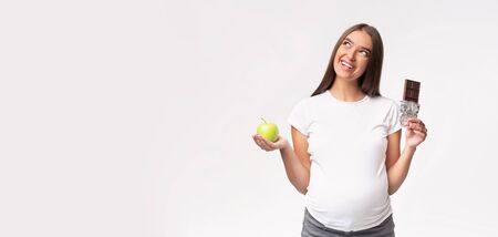 Pregnant Lady Holding Apple And Chocolate Choosing Between Healthy And Unhealthy Products Standing Over Gray Studio Background. Panorama, Copy Spaceの写真素材