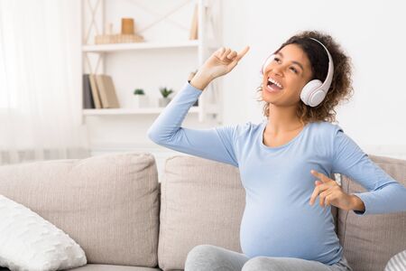 Positive pregnant woman enjoying music in headphones, relaxing on sofa at homeの写真素材
