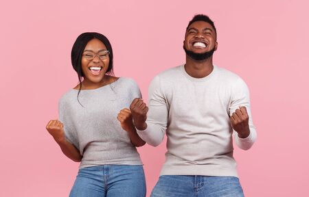 Young black emotional partners rejoice success, clench fists and exclaim with triumph, standing next...