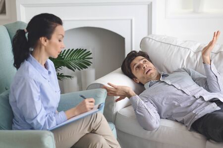 Puzzled man laying on coach at psychologist cabinet, sharing thoughts with specialist, free space