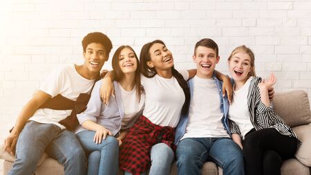 Happy teenagers embracing, sitting on sofa and smiling together to camera over light wallの写真素材