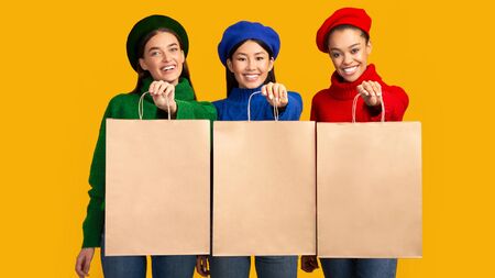 Shopping. Three Multiethnic Girls Holding Blank Shopper Bags Smiling At Camera Standing On Yellow Studio Background. Mockup, Panoramaの写真素材