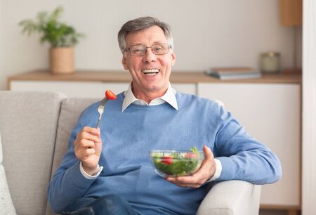 Nutrition in older age. Elderly man eating vegetable salad smiling sitting on sofa at home. Selective focusの写真素材