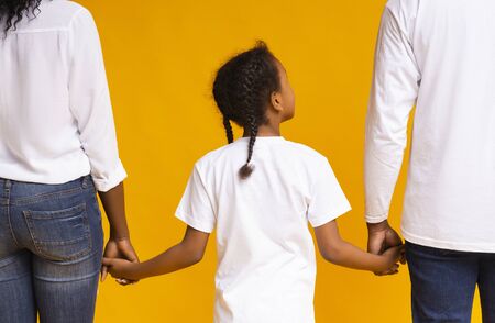 Family connection. Rear view of african mother, father and daughter holding hands together, yellow studio background, closeupの写真素材