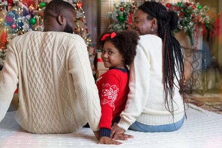Cute little afro girl looking at camera over shoulder while celebrating Christmas with her parents at homeの写真素材