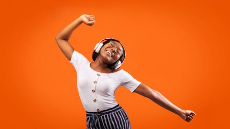 Happy African American Girl In Wireless Headphones Listening To Music Dancing In Studio Over Orange Background. Panoramaの写真素材