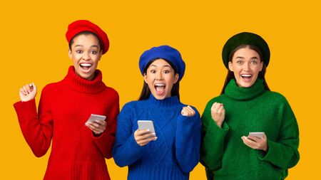 Three Excited Diverse Girls Holding Smartphones And Gesturing Yes Celebrating Success Standing Over Yellow Studio Background. Panoramaの写真素材