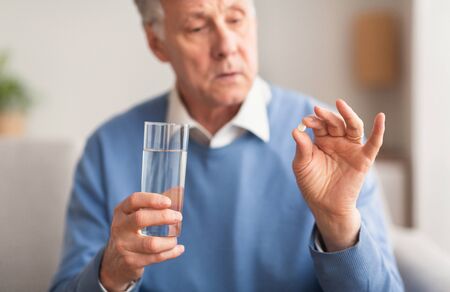 Medication Therapy. Senior Man Holding A Pill And Glass Of Water Sitting On Sofa At Home, Selective Focusの写真素材
