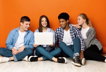 Diverse students looking at laptop, watching videos in the Internet, sitting on floor over orange...