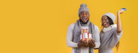 We are rich. Happy black couple with Christmas gifts and credit card over yellow background,...