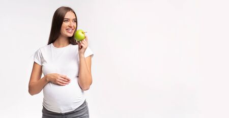 Pregnancy And Healthy Nutrition. Expectant Girl Eating Apple Touching Belly Standing On White Studio Background. Panorama With Free Spaceの写真素材