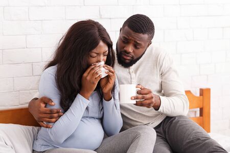 Handsome afro man taking care about his sick pregnant woman at home, giving her hot drink, copy spaceの写真素材